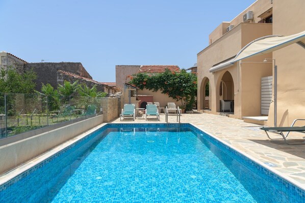A swimming pool with lounge chairs, a covered seating area, and a view of a historic building in the background. - Villa Nephele, 3 BDR, Forest Views, Private Pool, Furnished Terrace, Crete (Rethymno)