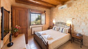 A bedroom with a stone wall, wooden ceiling beams, a bed with white linens, a bedside table with a vase of flowers, and a window with a view of greenery. - Villa Dione, 3 BDR, Forest Views, Private Pool, Furnished Terrace, Crete (Rethymno)