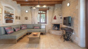 A cozy living room with a stone wall, wooden beams, a sofa, a television, and a fireplace. - Villa Dione, 3 BDR, Forest Views, Private Pool, Furnished Terrace, Crete (Rethymno)