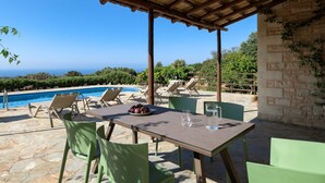 A covered outdoor area with a pool, lounge chairs, and a table with a fruit bowl and glasses. - Villa Dione, 3 BDR, Forest Views, Private Pool, Furnished Terrace, Crete (Rethymno)