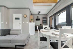 A modern living space with a sofa, dining table, chairs, a kitchen area, and large windows.