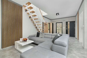 A modern living room with a grey sectional sofa, a white coffee table, and a wooden slatted wall. A white staircase with wooden steps leads to another room.