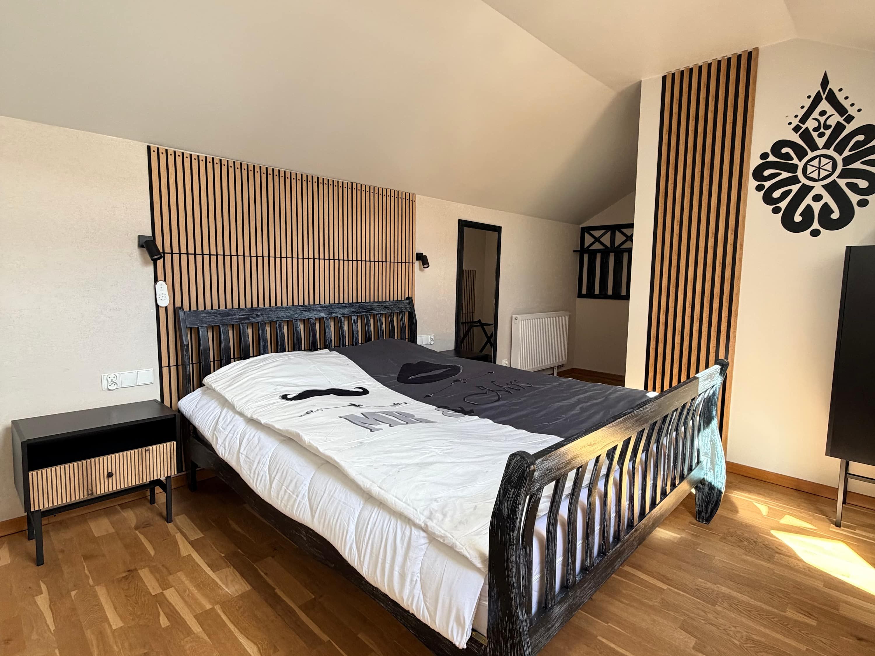 A bedroom with a wooden headboard, a bed with a white and black patterned bedspread, a bedside table, and a wooden floor.