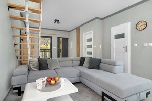 A modern living room with a grey sectional sofa, a white coffee table with apples and a box, a wooden ladder, and a clock on the wall.