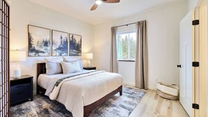Une chambre à coucher comprenant un lit, des tables de chevet, un ventilateur de plafond, une fenêtre avec des rideaux et des œuvres d’art accrochées au mur.