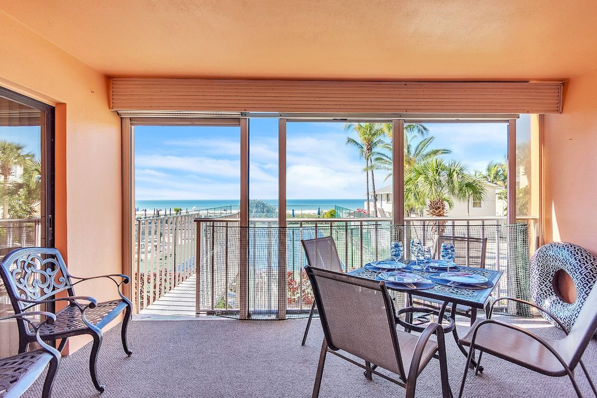 A balcony with a table and chairs, overlooking a beach and palm trees.