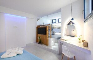 A modern, minimalist room with a bed, a small seating area, a desk, and a kitchen in the background. - Villa Evadne, 2 BDR, Outdoor Pool, Indoor Hot Tub, Furnished Terrace, Santorini (Santorini)