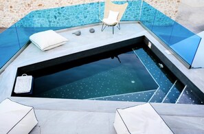 A hot tub with a stone wall backdrop, a white chair, and a small table with a white cushion. - Villa Evadne, 2 BDR, Outdoor Pool, Indoor Hot Tub, Furnished Terrace, Santorini (Santorini)