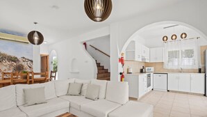 Uma sala de estar moderna com um sofá seccional branco, mesa de centro de madeira e uma escada que leva a uma cozinha com armários brancos e painel de azulejos.
