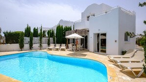 Uma piscina com espreguiçadeiras e uma mesa, cercada por um edifício branco com varanda.