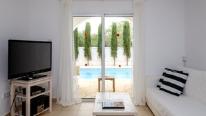 Sala de estar com televisão de tela plana em suporte, sofá com almofadas listradas e vista para uma piscina através de portas de vidro.