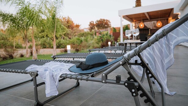 A sunbed with a hat and a towel, a dining area with tables and chairs, and a building with a chimney in the background. - Villa Hydraea, Garden Views, 2 BDR, Ptivate Pool, Furnished Terrace, Soroni (Rhodes)