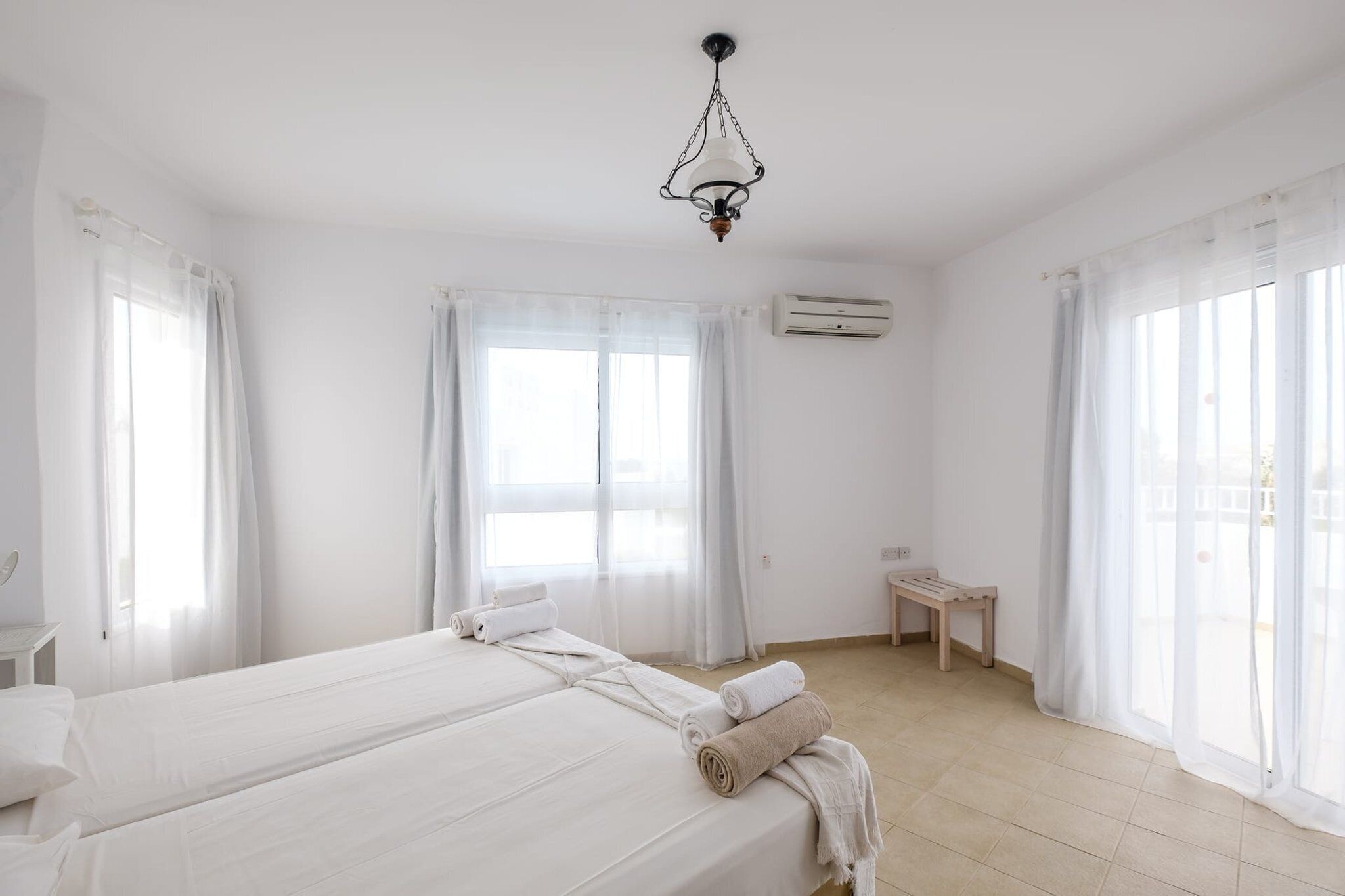 Una habitación de hotel luminosa y blanca con dos camas, una mesita pequeña y una lámpara en el techo.