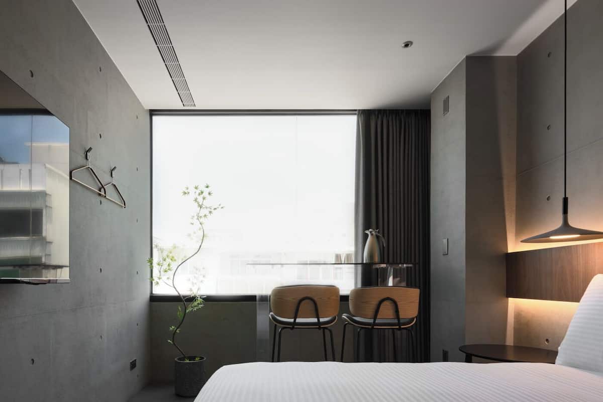Ein modernes Hotelzimmer mit einem großen Bett, zwei Sesseln, einem kleinen Tisch, einer Topfpflanze und einem großen Fenster mit Blick auf die Stadt.