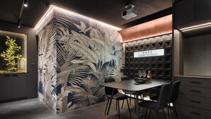 Une chambre d’hôtel moderne aux couleurs sombres, agrémentée d’une grande fresque murale, d’un coin repas avec une table et des chaises, et d’un mini-bar.