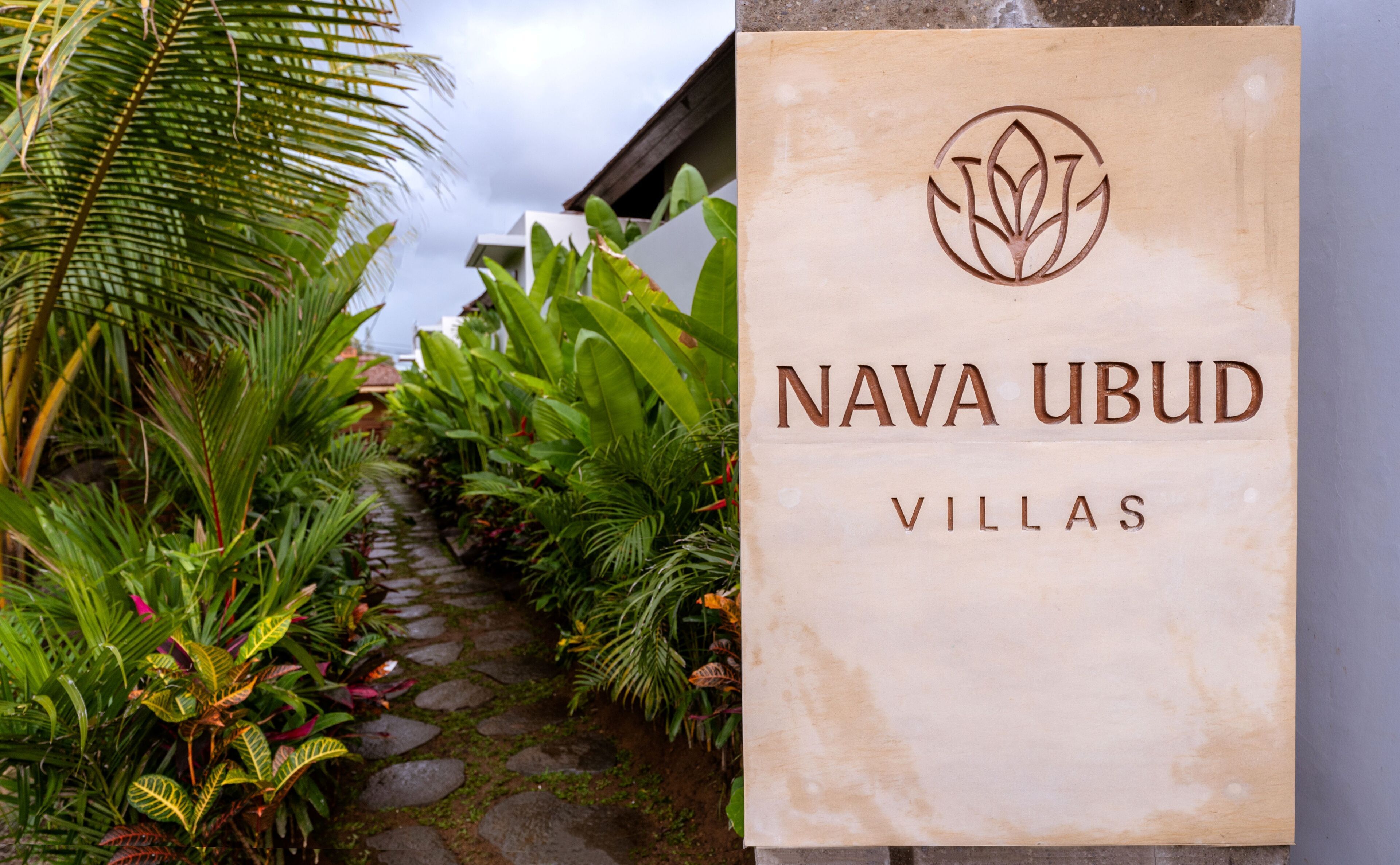 A sign for 'NAVA UBUD VILLAS' with a logo, set against a backdrop of lush greenery and a stone pathway.