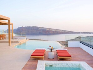 A rooftop infinity pool with wooden deck, two red lounge chairs, a small table with a drink, and a view of the sea and cliffs. - Villa Damaris, Divine Sunset & Sea Views, Private Pool, Furnished Terrace, Oia (Santorini)