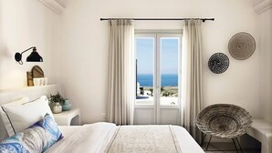 A bedroom with a large bed, a view of the sea, a wicker chair, and a wall-mounted lamp. - Thelia Suite, Divine Sunset & Sea Views, Private Jacuzzi, Furnished Terrace, Oia (Santorini)