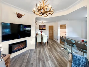 Uma sala de estar moderna com lareira, uma televisão de tela plana grande, uma área de jantar com mesa e cadeiras, e uma cozinha com balcão.