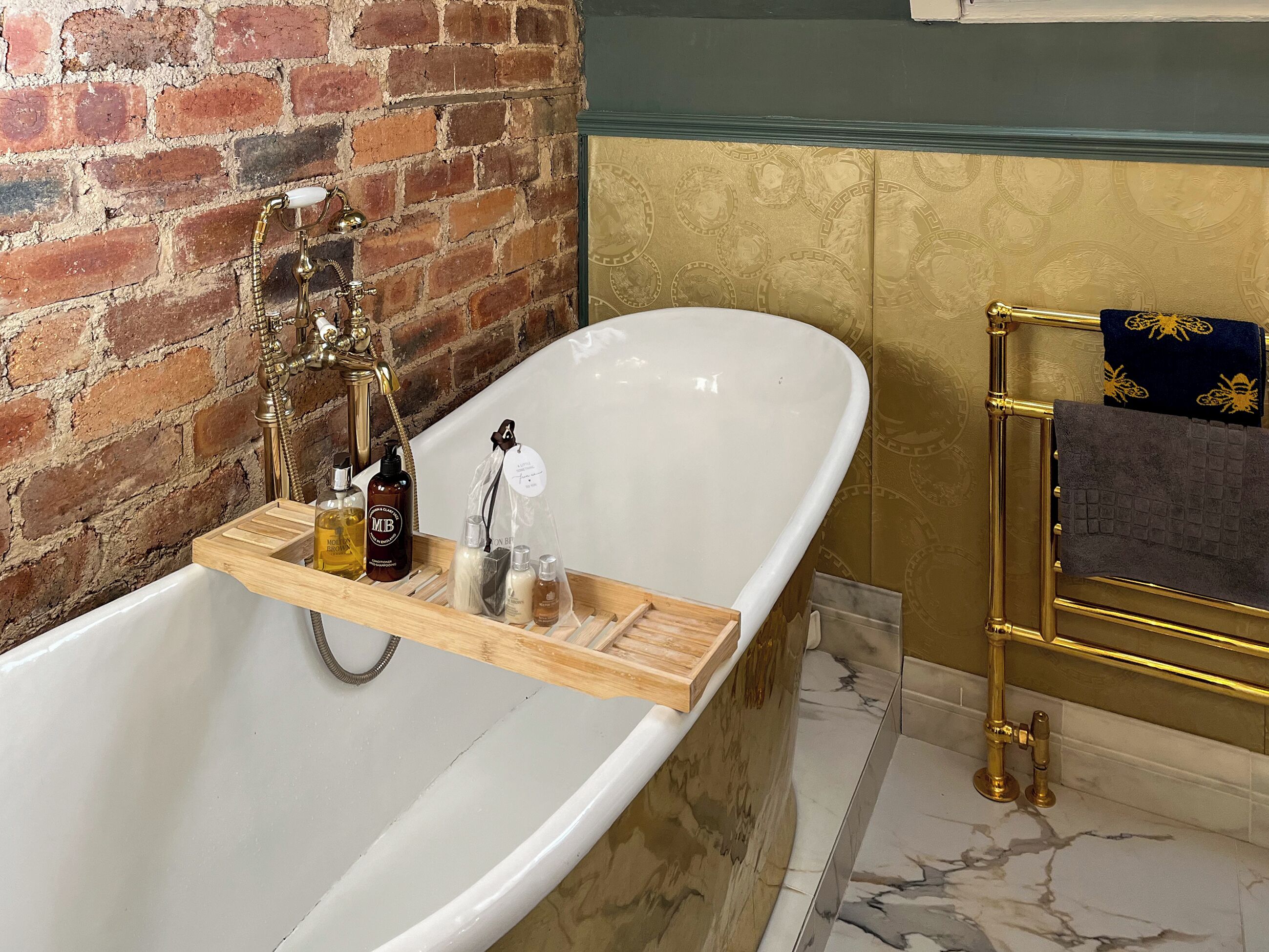 A bathroom with a freestanding bathtub, a wooden shelf with toiletries, a gold towel rack, and a brick wall.