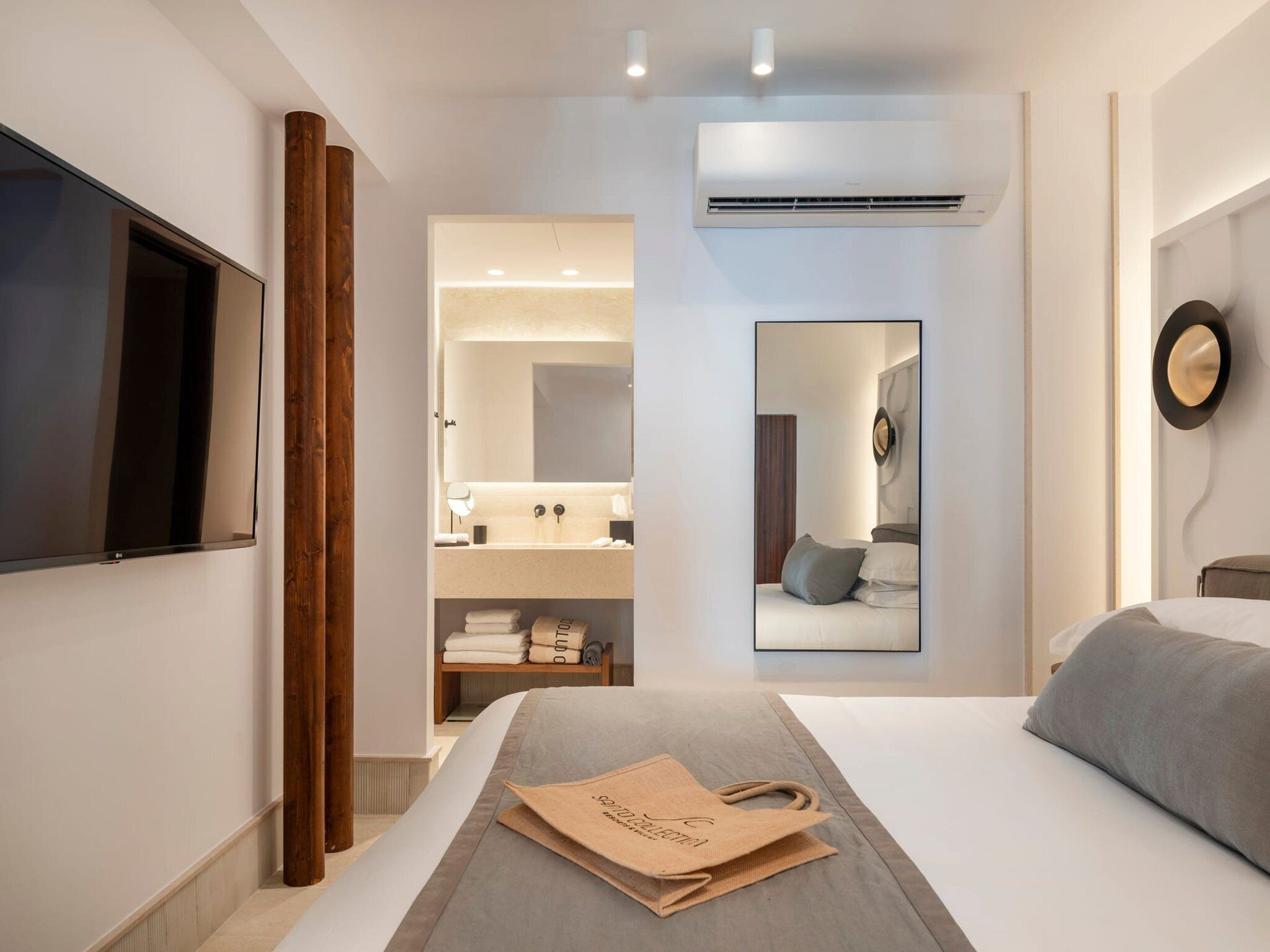 A modern hotel room with a large bed, a flat-screen TV, a bathroom with a mirror, and an air conditioning unit.