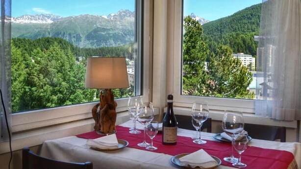 A table set for dining with a bottle of wine, glasses, and a lamp, overlooking a mountainous landscape. - Mountain Loft - DuLac 33 (Sankt Moritz)