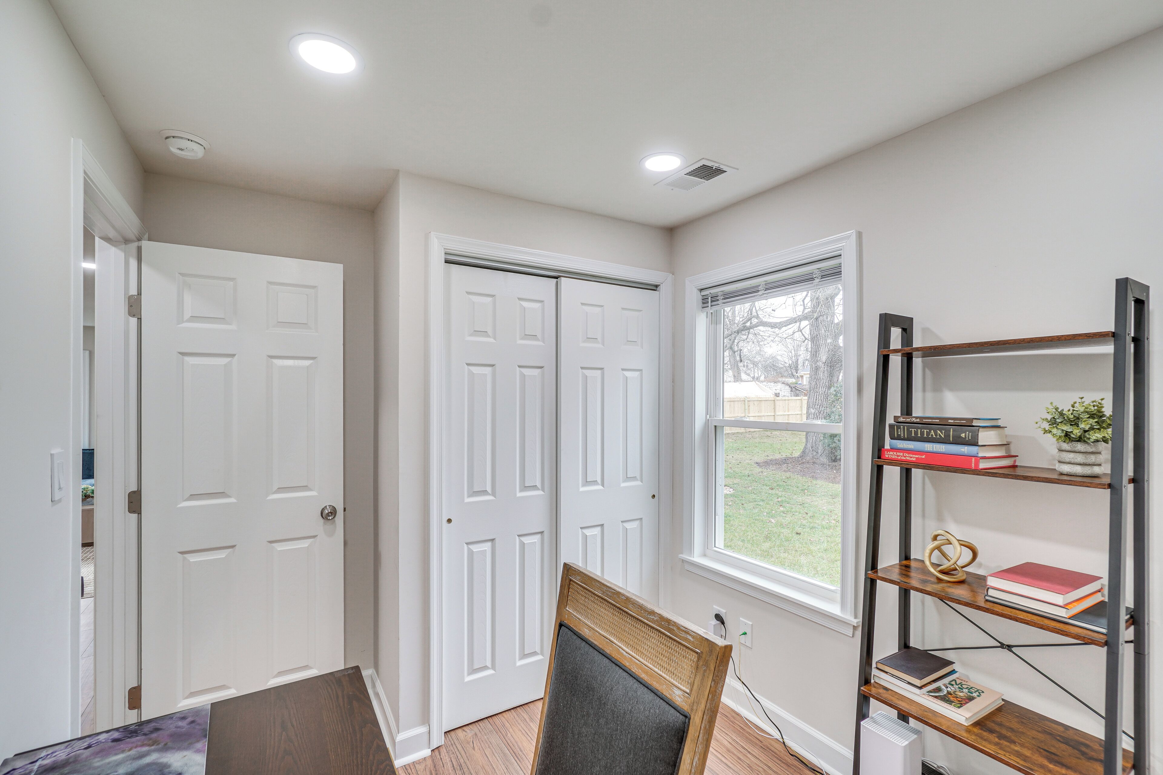 A room with white doors, a window with a view of a yard, and a bookshelf with books and a plant.
