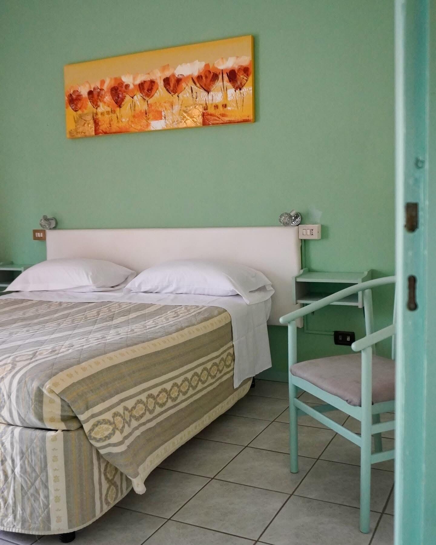Un dormitorio con cama, almohadas, una silla, mesita de noche, un reloj de pared y un cuadro.