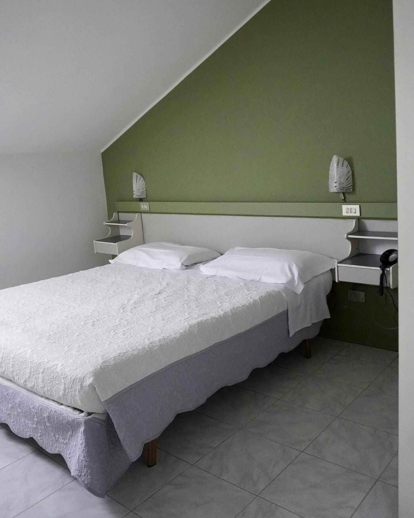 A single bed with a white bedspread and pillows, a bedside table with a lamp, and a telephone on a green wall.