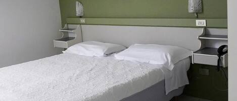 A single bed with a white bedspread and pillows, a bedside table with a lamp, and a telephone on a green wall.