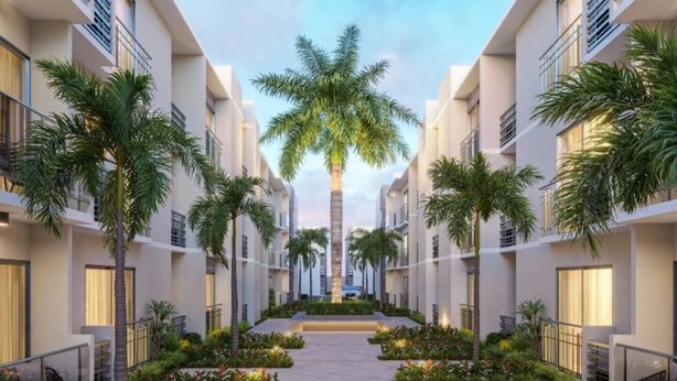 A modern hotel corridor with a central pathway flanked by palm trees and plants, leading to a building with balconies and large windows. - Moderno Apto en B Varo Island Piscina Playa (Punta Cana)