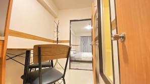 Ein schlichter Raum mit einem Holztisch, einem schwarzen Stuhl, einem Bett mit weißer Matratze und einer Holztür.