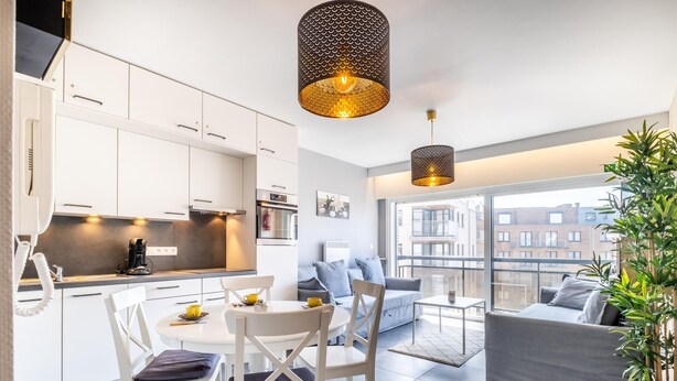 Um apartamento moderno com cozinha, sala de jantar e sala de estar. Possui uma janela grande com vista para a cidade, uma mesa de jantar com cadeiras e um sofá.