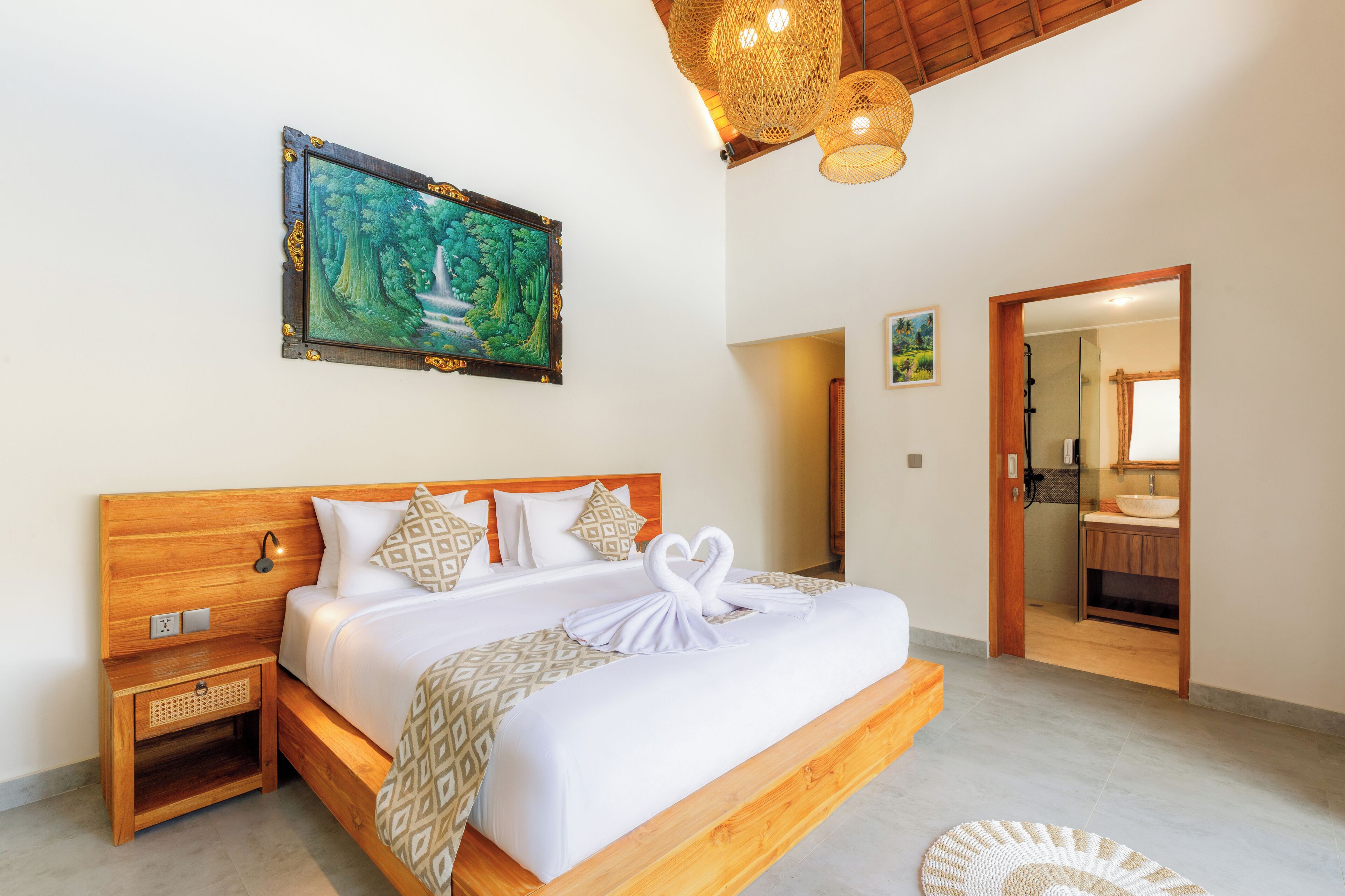 A spacious bedroom with a large bed, a wooden headboard, a painting of a forest, and a bathroom visible through an open door.