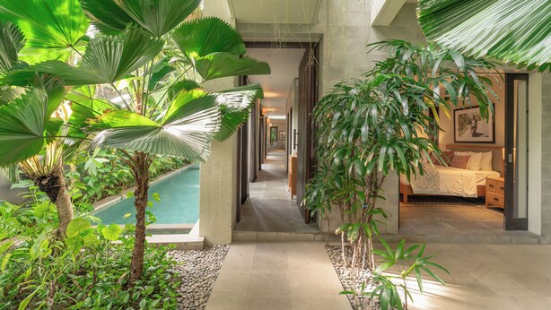 A hotel corridor with a pool, indoor plants, and a bedroom with a bed and nightstand. - Villa Ixora by Alfred in Bali (Canggu)