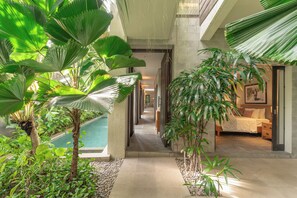 A hotel corridor with a pool, indoor plants, and a bedroom with a bed and nightstand. - Villa Ixora by Alfred in Bali (Canggu)