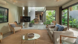 A modern living room with a glass coffee table, sofas, and a TV. There is a staircase leading to an upper level, and large windows with a view of greenery outside. - Villa Ixora by Alfred in Bali (Canggu)