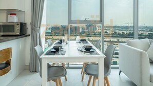 A modern dining area with a white table set for four, grey chairs, a white sofa, and a large window offering a city view. - Zapbed-1 BR in District One Residence 16 (Dubai)