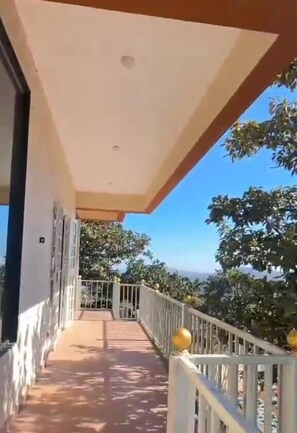 A covered balcony with a railing, a clear view of trees and a distant landscape. - Kedareshwar Agro Tourism (Wai)