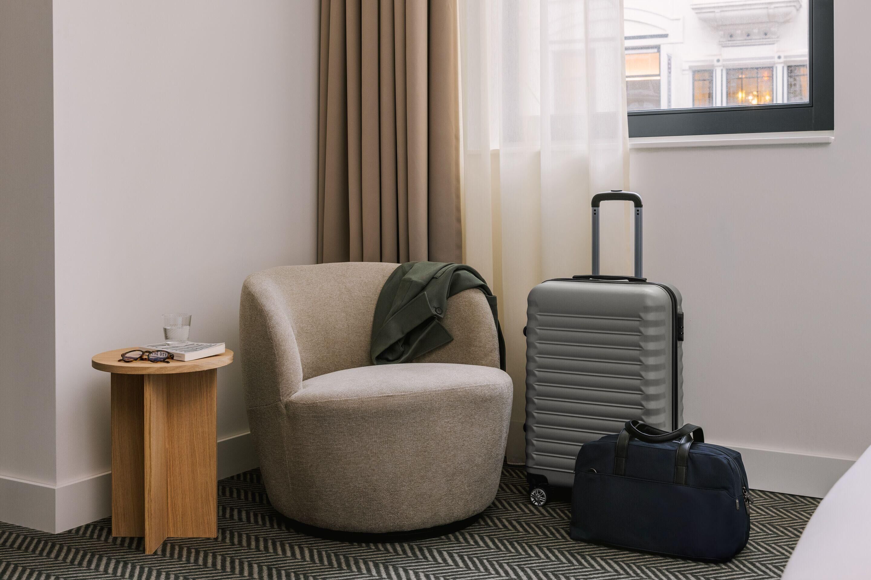 A hotel room with a single armchair, a small wooden side table, a grey suitcase, and a dark blue duffel bag.