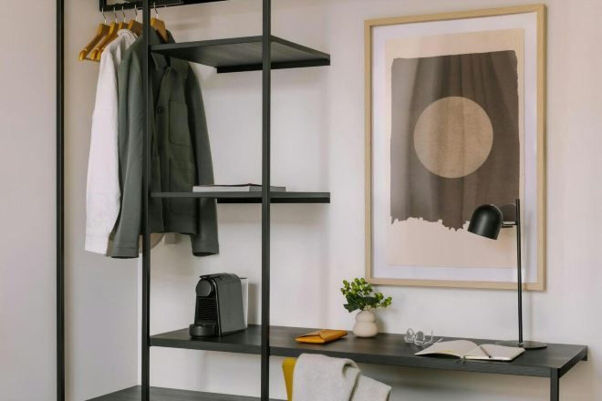 A modern room with a black metal shelving unit, a framed abstract painting, a desk with a lamp, and a small potted plant.