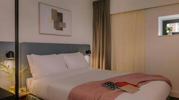 A hotel room with a bed, a nightstand, a wall-mounted lamp, a window with curtains, and a framed picture on the wall.