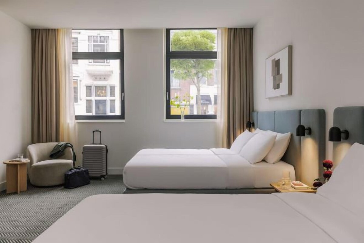 A hotel room with a bed, a chair, a suitcase, and a view of buildings and trees through the windows.