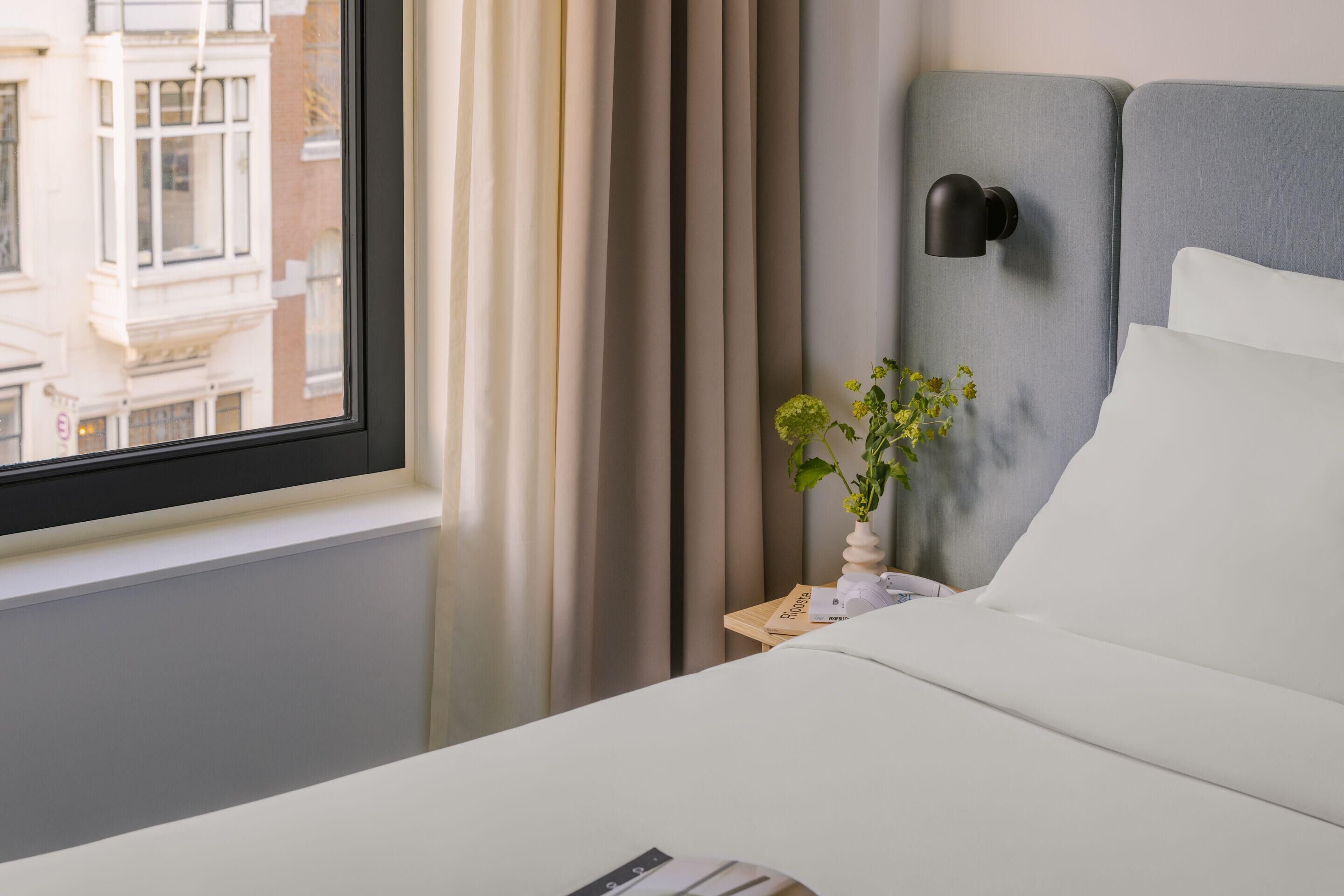 A hotel room with a bed, a bedside table with a lamp, a window with curtains, and a view of buildings outside.