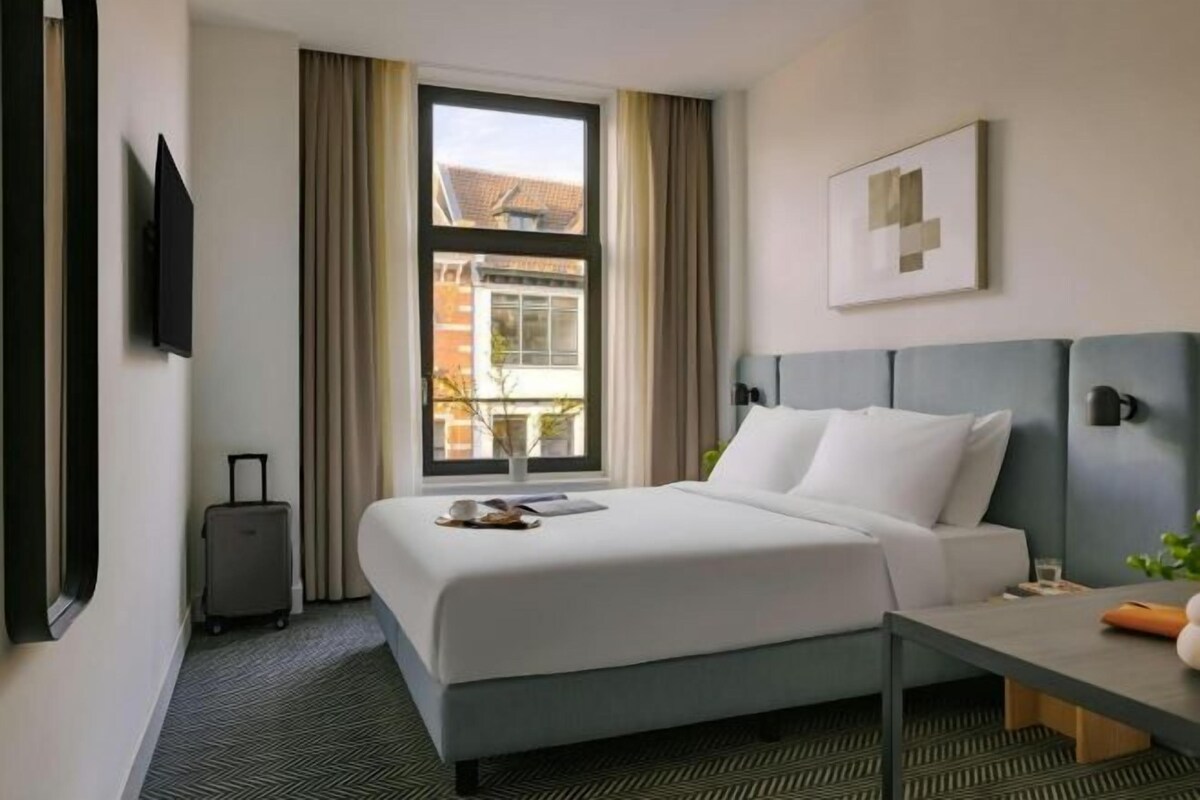 A hotel room with a bed, a desk, a chair, a TV, a window with curtains, and a view of buildings outside.