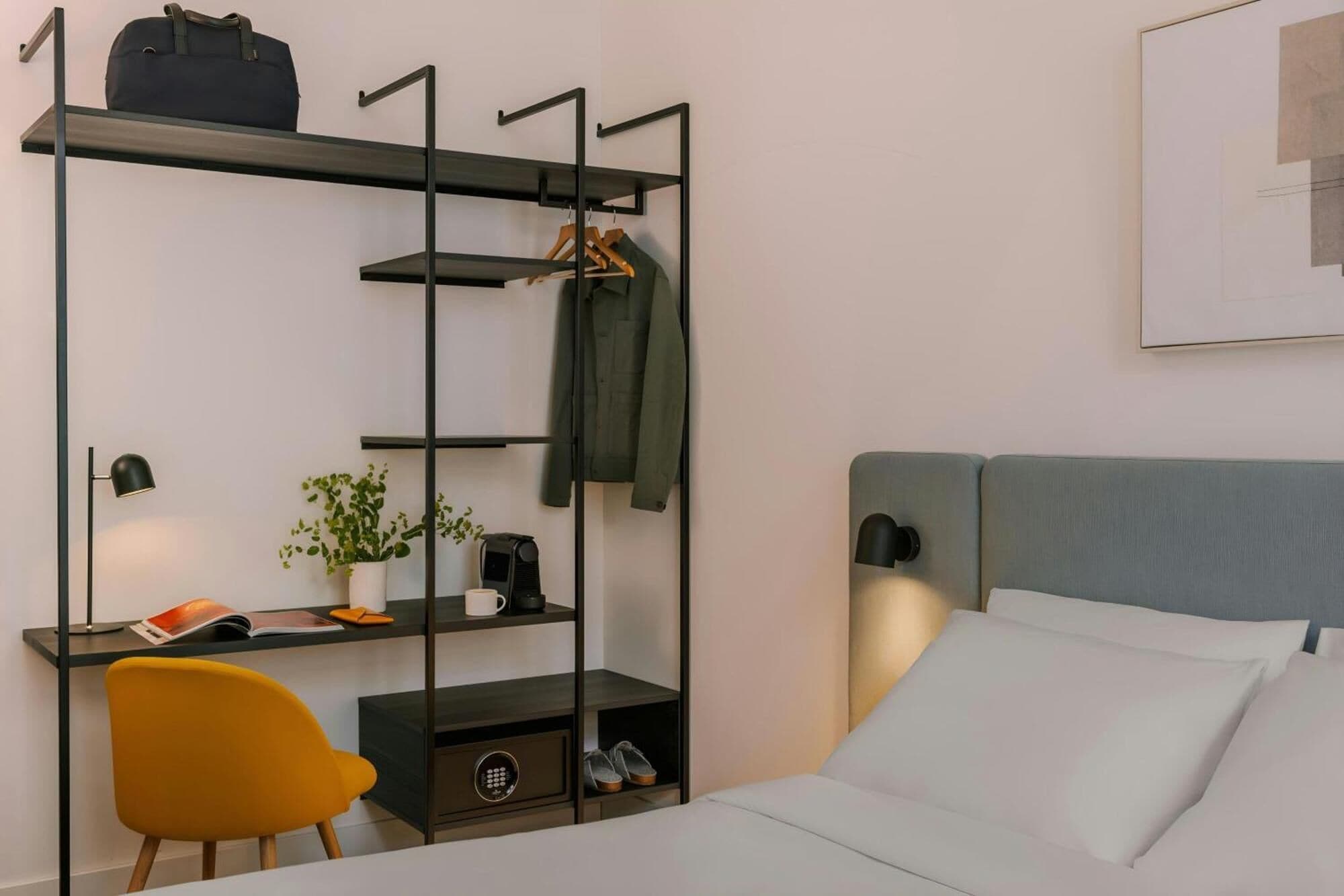 Ein Hotelzimmer mit einem Bett, einem Schreibtisch mit Stuhl, einer Regaleinheit mit Jacke und Pflanze sowie einer Wandlampe.
