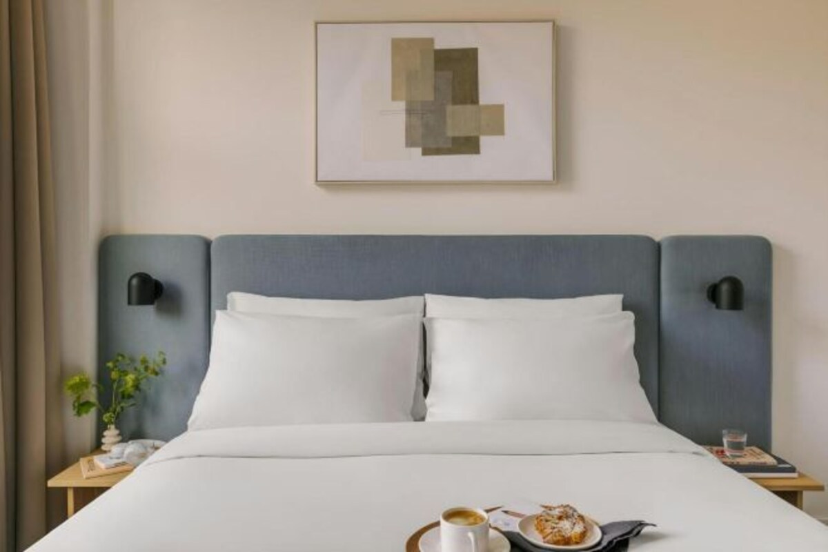 A hotel room with a bed, two bedside tables, a wall-mounted artwork, and a small plant on the left side.
