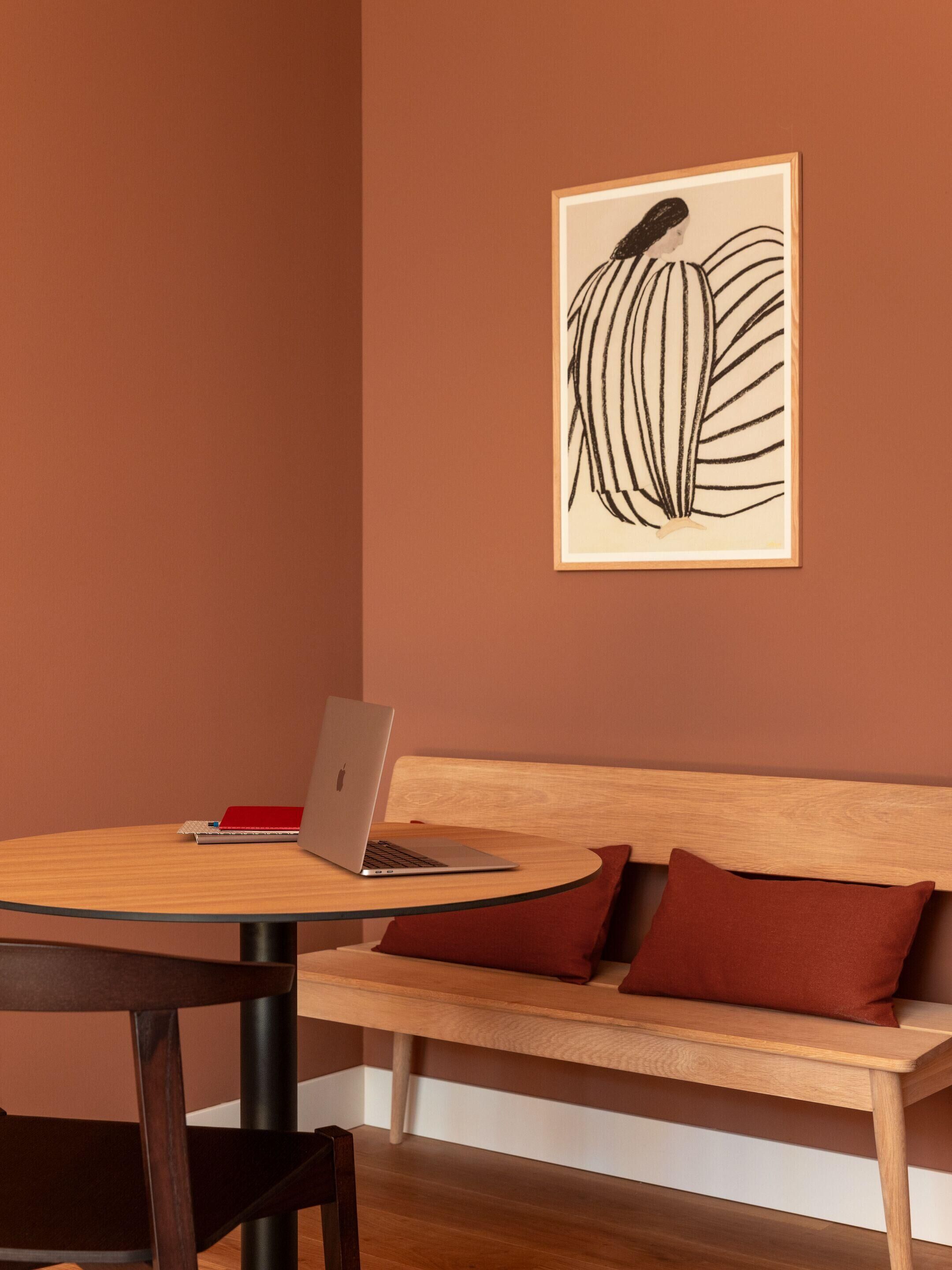 A room with a wooden table, a laptop, a red notebook, and a wooden bench with red cushions.