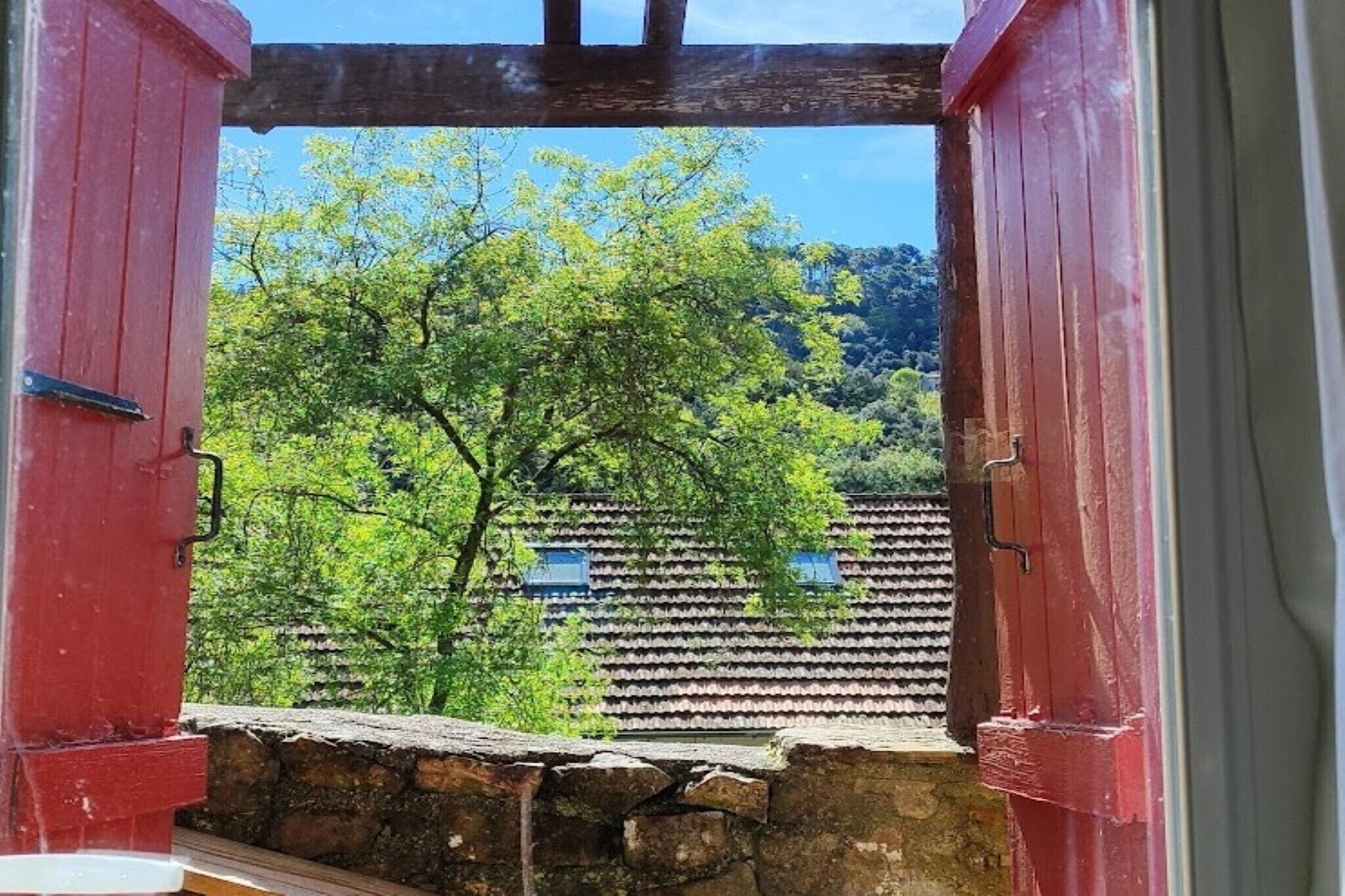 Vista attraverso una finestra con cornice in legno e persiane rosse, su un edificio con tetto in tegole e alberi verdi e rigogliosi.