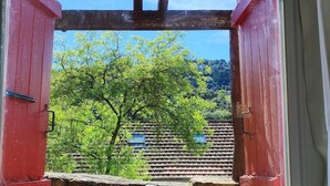 Udsigt gennem et vindue med træramme og røde skodder, der viser et hus med tagsten og frodige grønne træer.